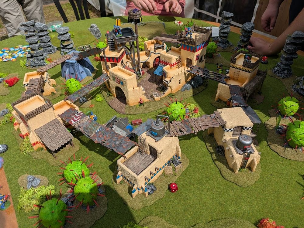 A game of Warhammer 40,000 in progress, featuring classic adobe Ork builings and spiky green cacti. 