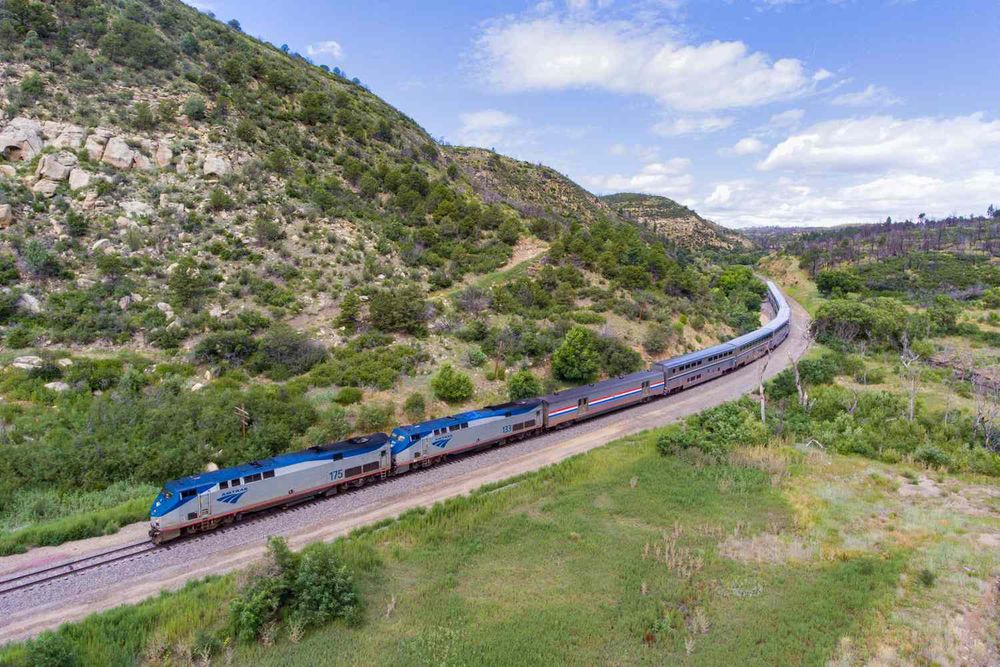 An Amtrak passenger train with two blue-and-silver locomotives and several silver cars winds through a green mountain valley under a bright blue sky with scattered clouds. The train curves along a single track bordered by grass, shrubs, and rocky hillsides dotted with trees.