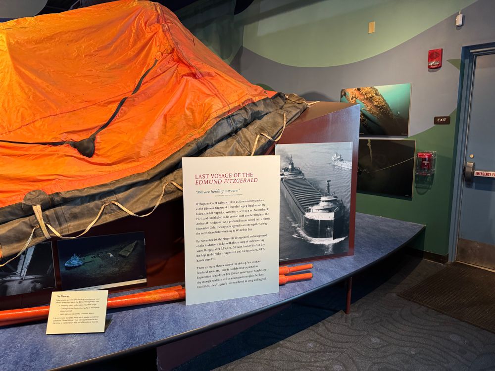 Museum display at the National Museum of the Great Lakes in Toledo featuring an orange life raft canopy from the Edmund Fitzgerald exhibit. A sign titled “Last Voyage of the Edmund Fitzgerald” describes the ship’s final moments and the loss of all 29 crew members. Historic photos of the freighter and underwater wreck images are displayed behind it.