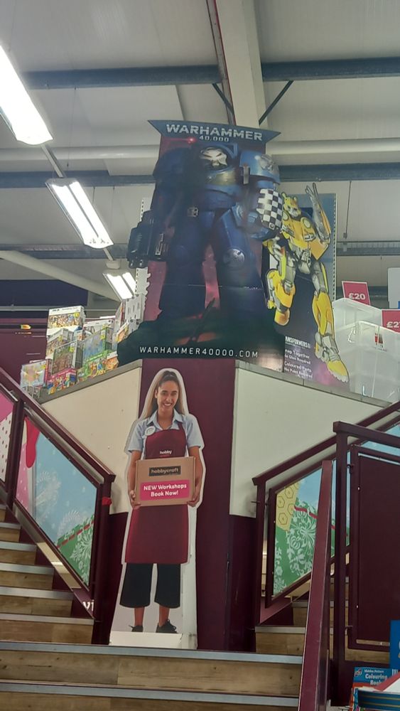 Cardboard standee of a Space Marine Terminator from Warhammer 40,000 standing on a display above two sets of stairs in a Hobbycraft. To it's right is a standee of a Transformer, below is a standee of a woman wearing a Hobbycraft apron carry a box that reads "NEW workshops book now!"
