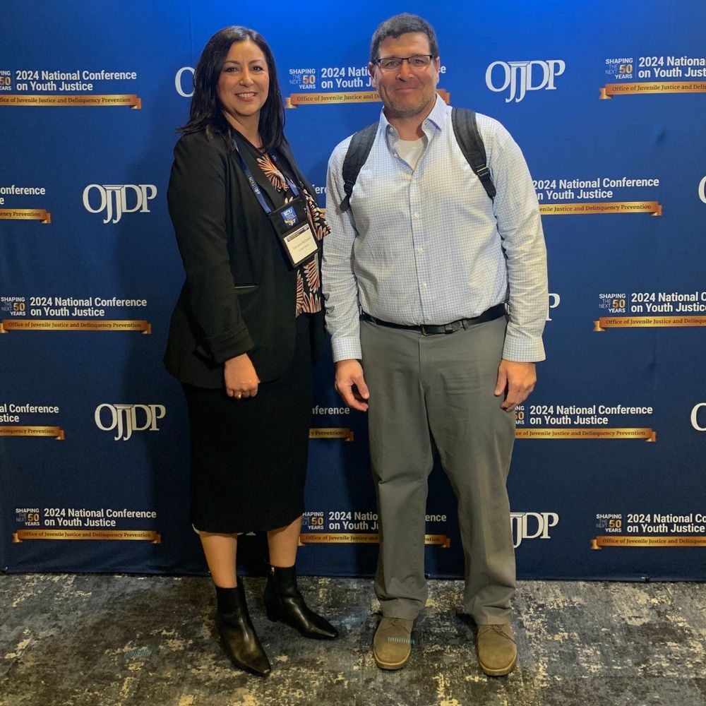 Director of Justice Georgina Mendoza McDowell and Director of Analytics Chris Scharenbroch stand in front of a backdrop announcing the 2024 OJJDP National Conference on Youth Justice.