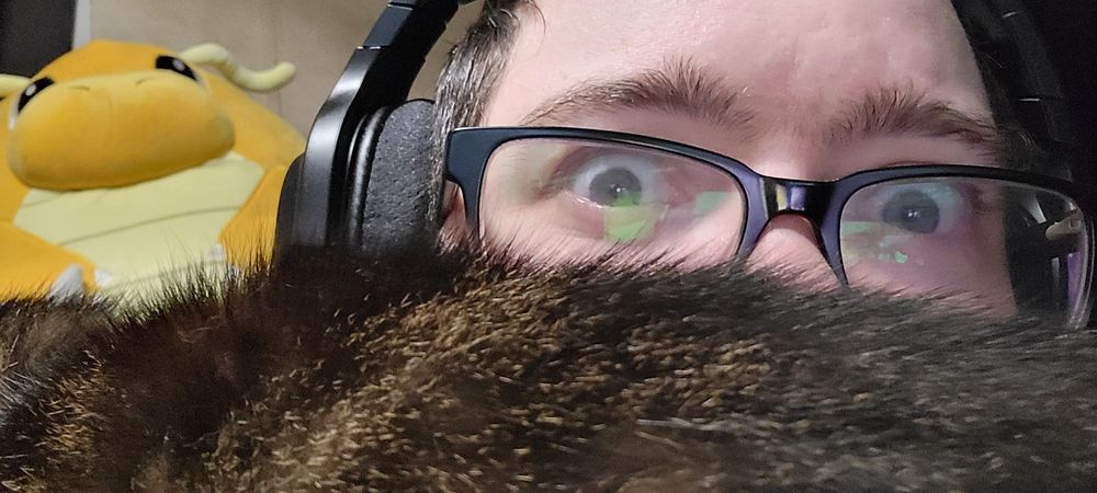 Me looking shocked with the lower half of my face covered by a cat laying across my chest. A dragonite squishmellow is visible over my shoulder.