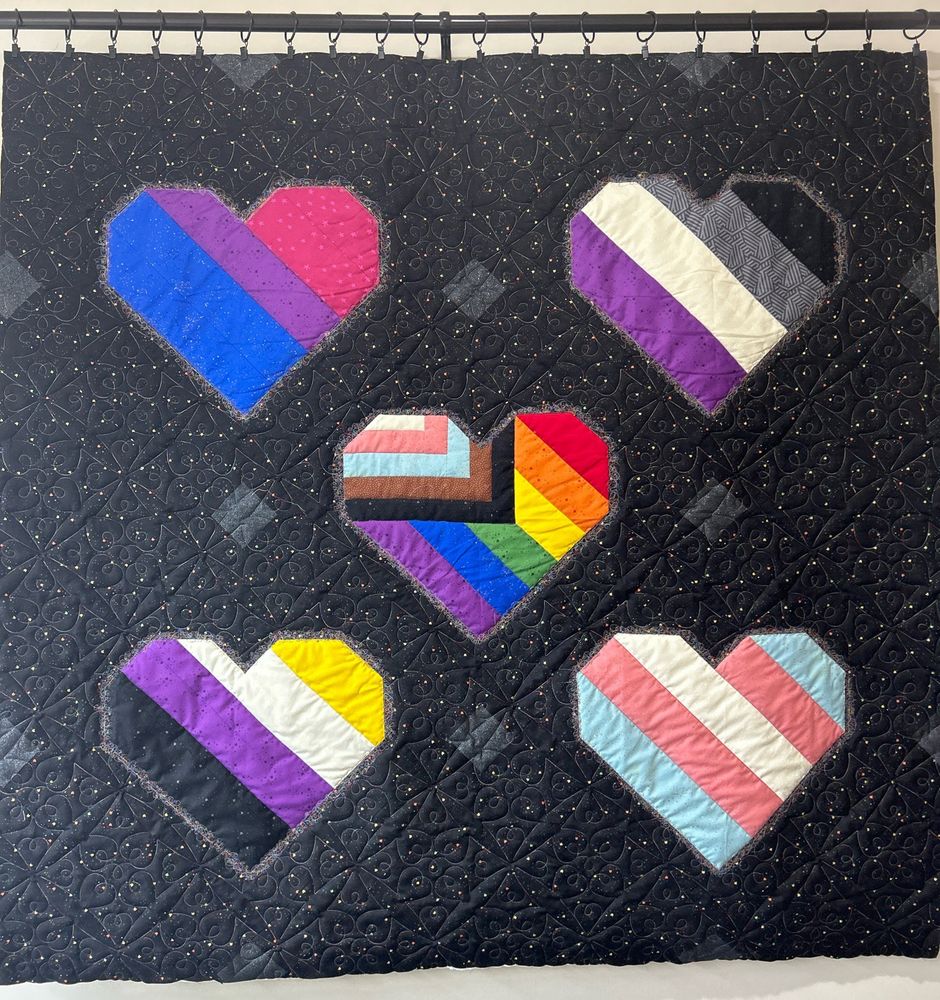 A square quilt hanging from a curtain rod. It has a black rainbow speckled background and features five hearts that showcase five Pride flags: the bisexual flag, the asexual flag, the progress pride flag, the nonbinary pride flag, and the trans pride flag. 
