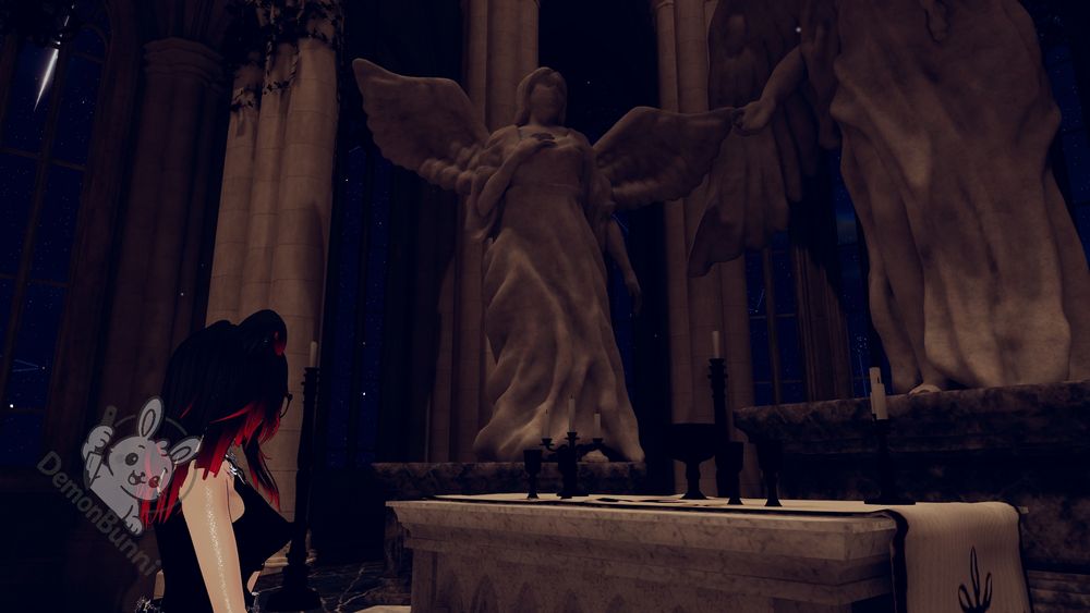 Photograph of a virtual church interior with a statue of an angel and a person. The statue is in the center, with large wings and a draped robe. A person with black and red hair stands to the left, facing the statue. A logo for "DemonBunni" is in the lower left. The church has tall columns and arched windows showing a starry night sky. Candles and ornate objects are on a marble altar in front of the statue.