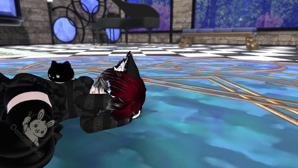 Photograph of a virtual avatar lying on a blue and gold patterned floor. The avatar has red and black hair, pointed ears, and is wearing a black outfit with a white stripe. A black cat plushie is next to the avatar. The avatar's outfit has a logo that says "DemonBunni" with a cartoon bunny holding a knife. In the background is a black piano, a bench, and windows.