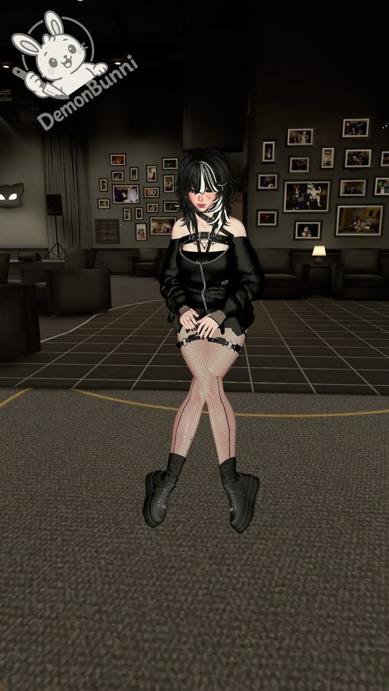 Photograph of a digital avatar with black and white hair wearing a black outfit and boots, sitting with legs crossed in a dark room. The avatar has a harness around their neck and chest, and fishnet stockings. Behind the avatar are dark couches and framed pictures on the wall. In the upper left corner is a logo with a white rabbit holding a knife, and the name "DemonBunni".