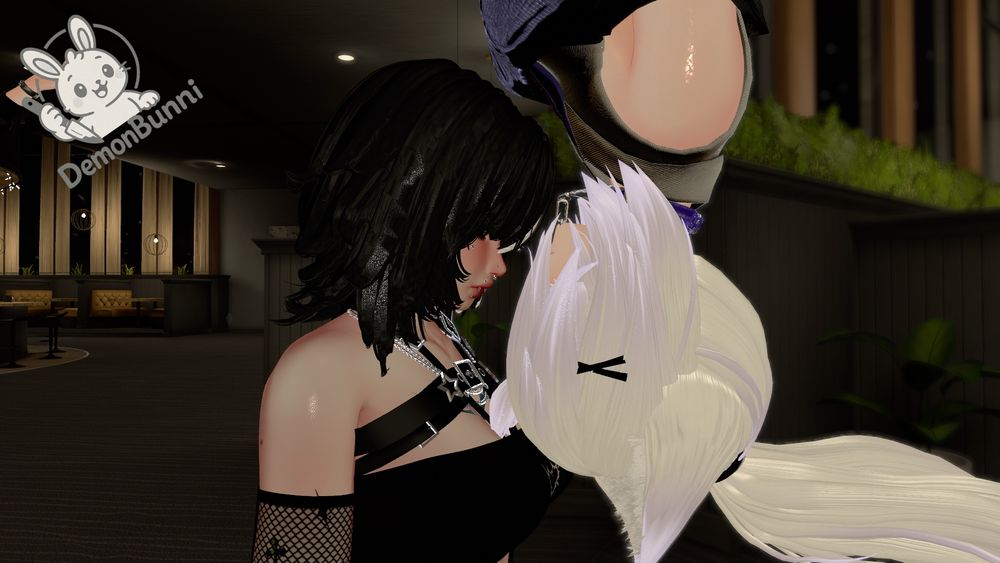 Photograph of two women embracing. One woman has long black hair, fair skin, and is wearing a black dress with a harness. The other woman has long white hair, fair skin, and is wearing a black and white outfit. The background is a dimly lit room with tables and chairs. The logo "DemonBunni" is in the upper left corner.
