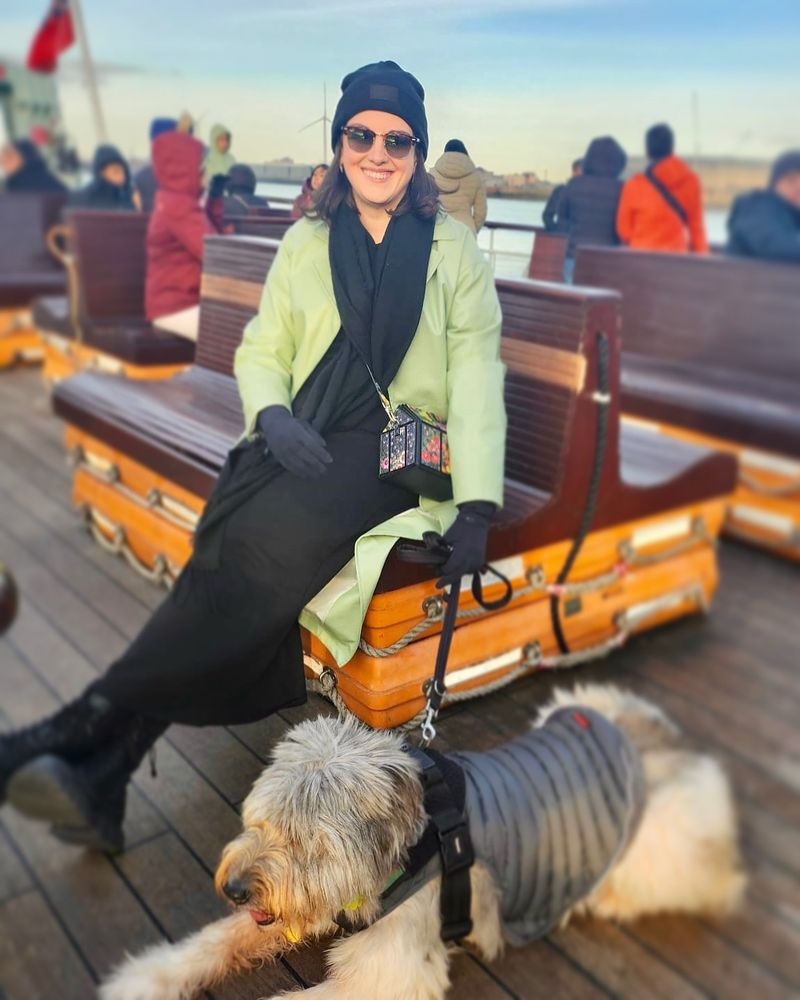 I am sat on a bench seat holding a leash. My dog Noodle is on the leash. She is lying down. She is a big grey and white fluffy shepherd dog. It is winter and we are both wearing coats.