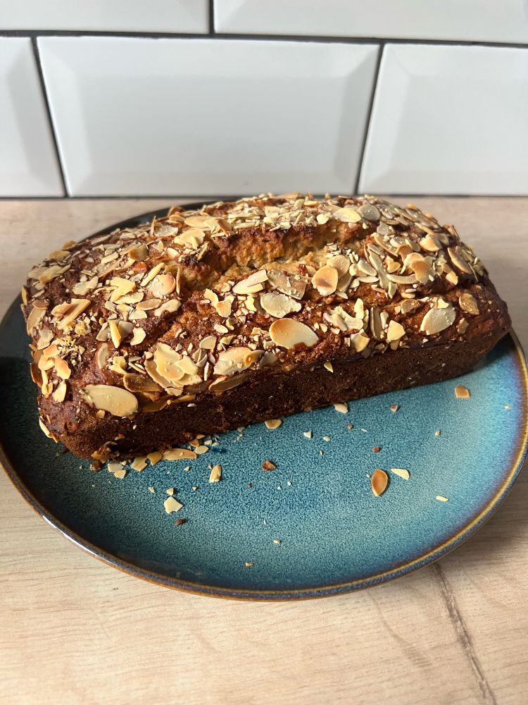 A photo of a loaf of banana bread. It is topped with flaked almonds. It is on a blue plate on a wooden worktop. 