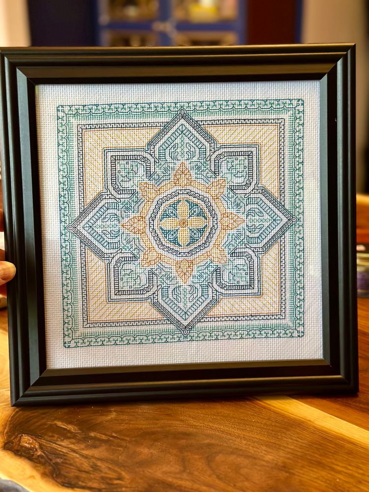 A framed black work embroidery piece in shades of green and yellow. There’s an eight pointed star or flower surrounded by a square multi layered border. It’s in a black wooden frame. 