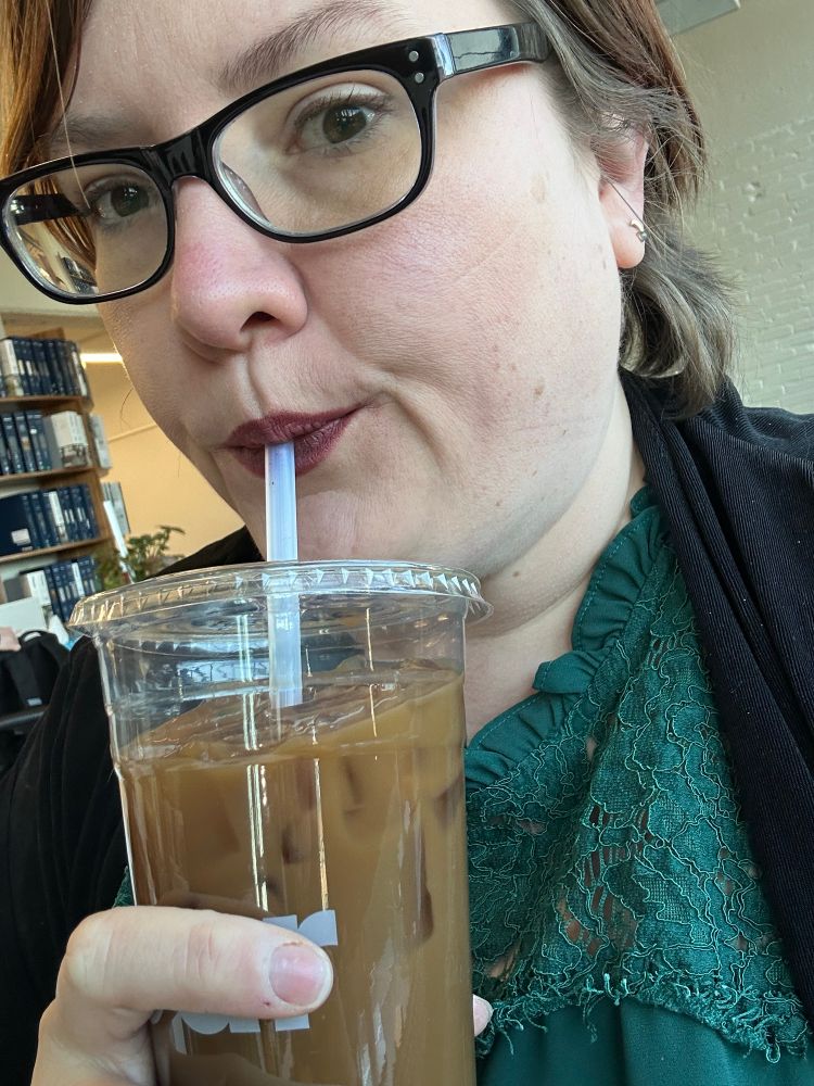 Selfie of a short haired white woman sipping cold brew from a straw. Her makeup is on point today, lipstick and a nice lil cat eye liner. 