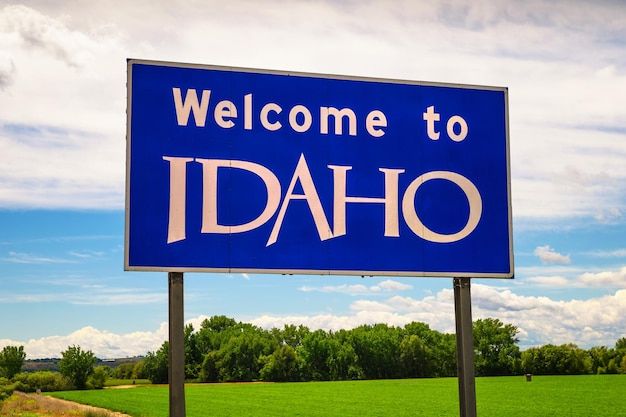 Welcome to Idaho sign. White text on a blue sign. It is outdoors with grass and a few trees and a sky with sunny but bored clouds in the background.