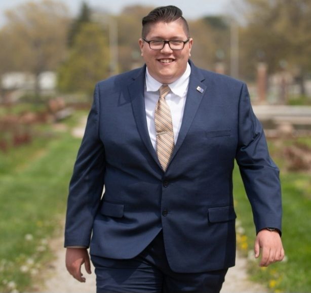 A portly young white male Republican in an ill-fitting suit walks down a rural road directly at the camera.