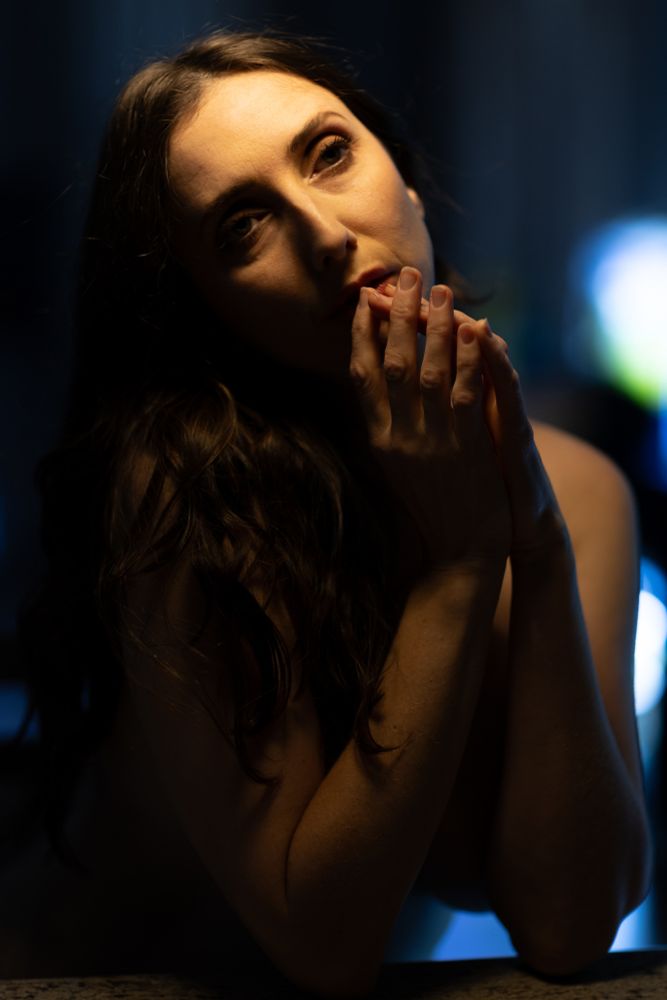 A woman sits leaning on a kitchen bar, nude with her arms clasped before her face, gazing off to the side. Light from a small light overhead lights her face and hair, the rest of her in shadow. With color from the sun leaking through closed curtains.