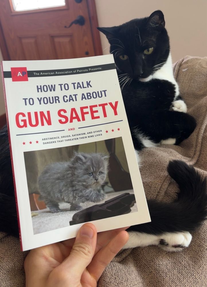me holding a book titled “How to talk to your cat about gun safety” in front of my cat