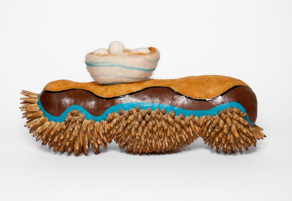 Ceramic sculpture shaped like a sea slug carrying a nest of eggs. The slug has many tiny, orange-brown legs and a graphic pattern of teal, brown, and mottled orange. The nest is needle-felted white with teal wisps and an orange interior. The nest has round, unglazed, porcelain eggs inside.
