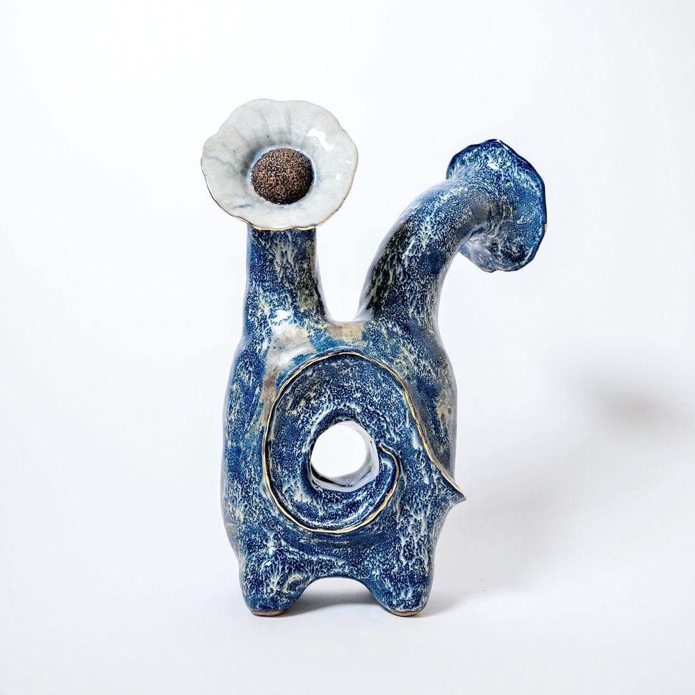An abstract sculpture that has four small feet (two visible), a “torso” with a golden swirl leading to a donut-style hole. It has two “necks” with flower “heads”. One flower faces away and one face-on.