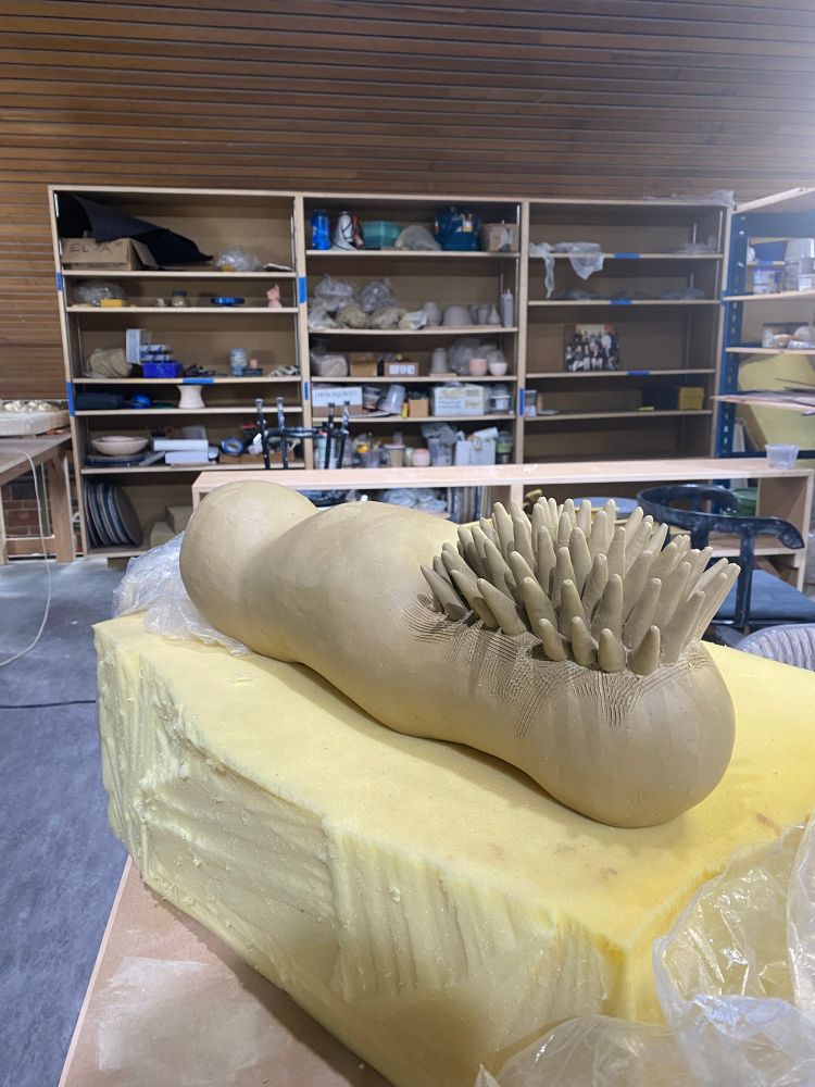 A clay sculpture of a worm-like creature with spikes partially covering. It is unfinished and sitting on foam in a ceramic studio.