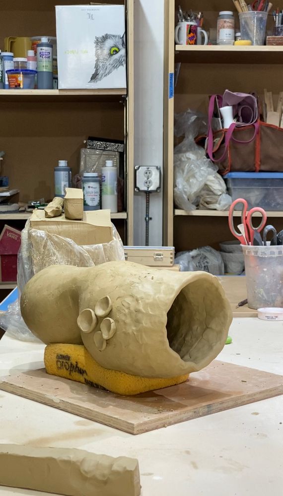 A hollow, unfinished sculpture of a worm sits atop a large sponge. The sculpture is made of clay and has temporarily placed suction cup-like appendages.