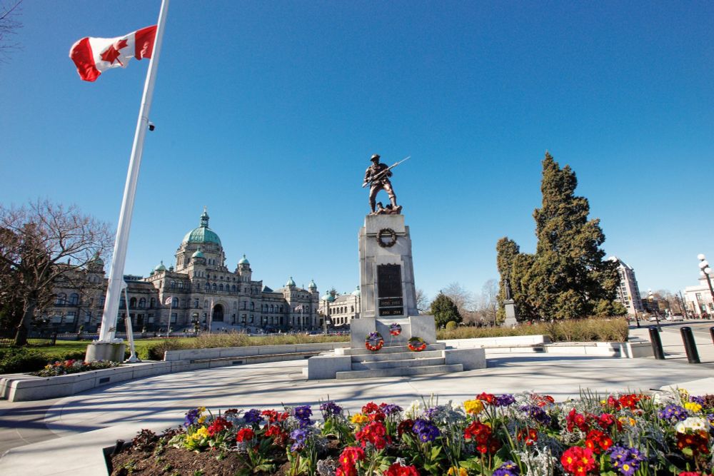 A Canadian flag at half-mast, in honor of Vimy Ridge day Apr. 9th, next to a war memorial statue of a soldier with a rifle, standing atop a stone pedestal adorned with wreaths. Colorful flowers are planted in the foreground. In the background, the historic British Columbia Parliament Buildings with a green-domed roof are visible under a clear blue sky.