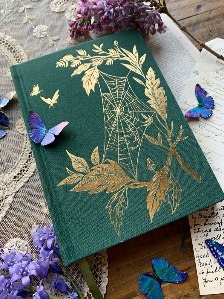A green linen bound notebook with a vintage looking gold foil stamped design of a spiderweb amongst the leaves 