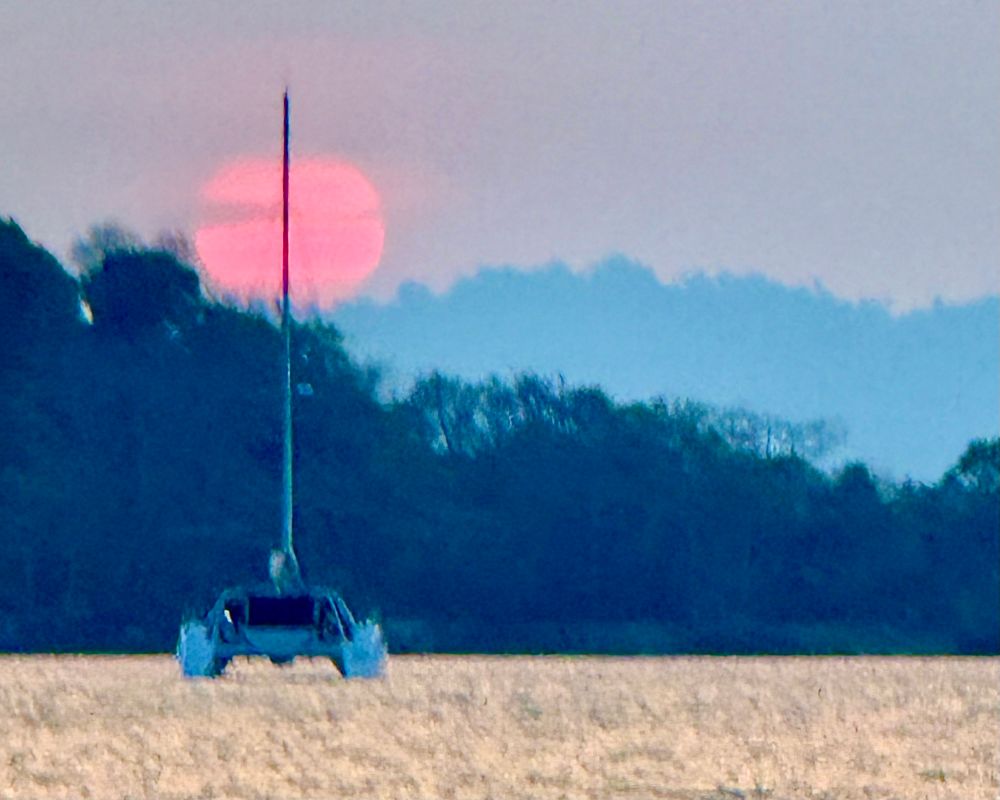 Setting sun behind bands of clouds and silhouette of tropical forest islands. In the foreground a catamaran yacht with the mast dividing the sun disk in two halves.