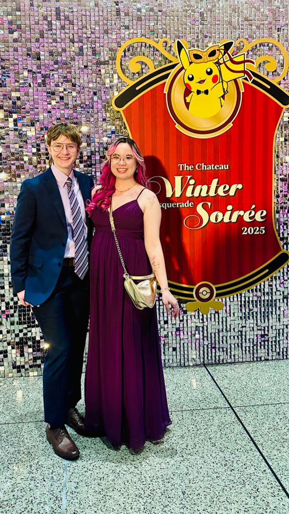 Myself and my fiancé Maddie standing in front of a silver sequin backdrop with a sign reading “Winter Masquerade Soirée 2025” next to me. I’m wearing a floor length purple dress and Maddie is wearing a navy blue suit with a striped purple tie and lavender shirt 