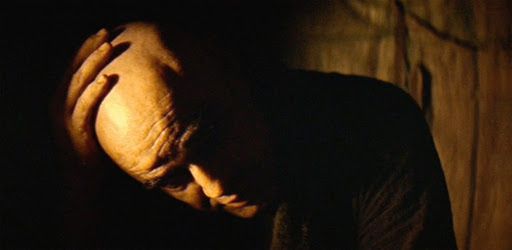 Col Kurtz from Apocalypse Now, played by Marlon Brando, from the extended final monologue of the film before he is murdered. His head is shaved, he sits in shadow with splashes of orange firelight, rubbing his scalp as he speaks.