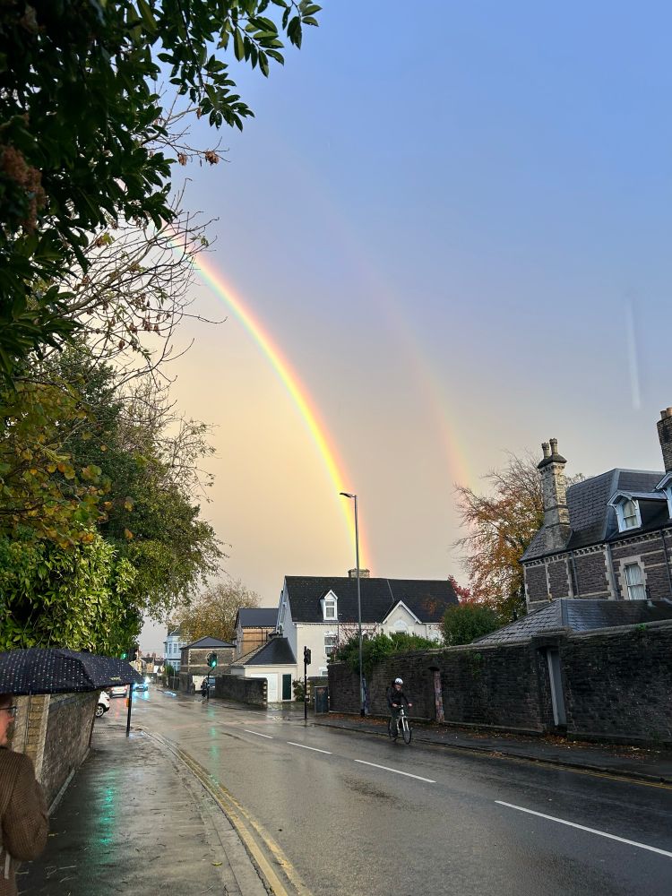 A picture of a deserted road after a rain shower. A double rainbow peeping through, one crescent appearing to emerge from a nearby chimney.