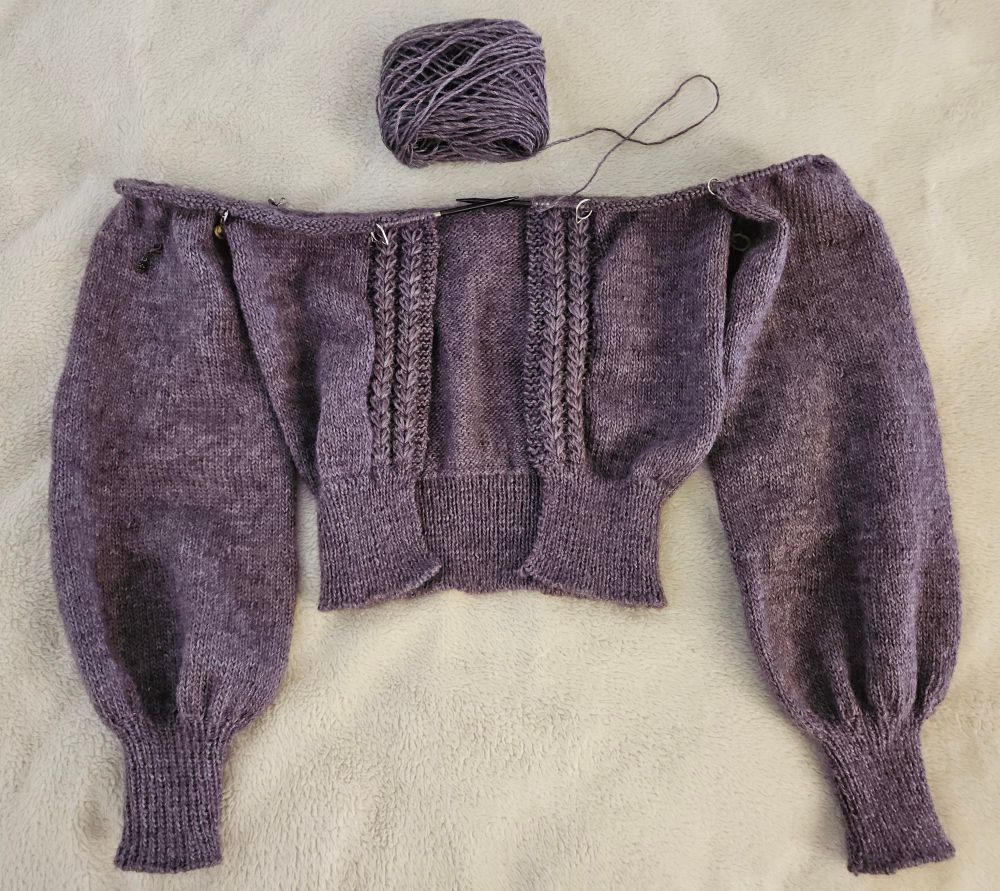 A purple hand-knit cardigan in progress lays flat on a white fuzzy background. The body and sleeves are on the cable of a knitting needle, awaiting the completion of the shoulder shaping and neckline.