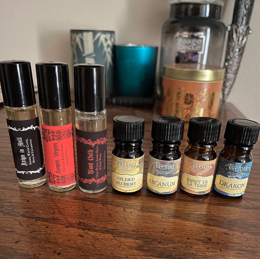Seven fragrance bottles sit on top of a wood dresser, all with black caps. There are 3 big ones and 4 small ones. The bigger ones’ labels vary in black, red, and white. The first one says “Reign in Hell - smoke, white clove, musk, cedarwood.” The second one says “Sanguis Virginis - metallic blood, black plums, & figs.” The third one says “Blood Oath - dried blood, amber resin, & rosewood.” The small bottles vary slightly in color, yellow, purple, orange, and blue, all with “Alchemia perfumes” logo. The first one says “Guilded Alchemy,” the second one says “Arcanum,” the third one says “Esprit de la Terre,” and the fourth one says “Drakon.”
Behind the seven fragrances are an array of candles and candle holders, and a small decorative dagger