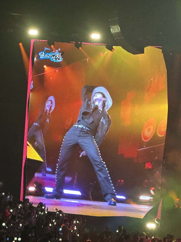 A Korean man on stage in a black cowboy outfit and brown hat striking a pose 