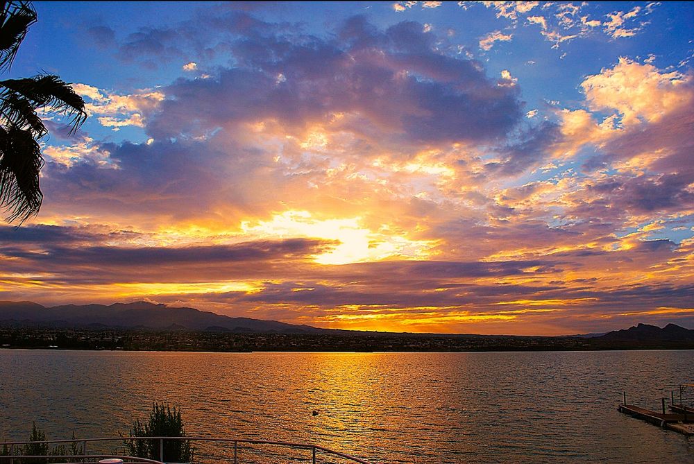 The sunrise from across the lake lights up the early morning clouds and sky in vivid colors with the lake taking on a golden hue. There’s bits of natural vegetation bleeding into the picture 