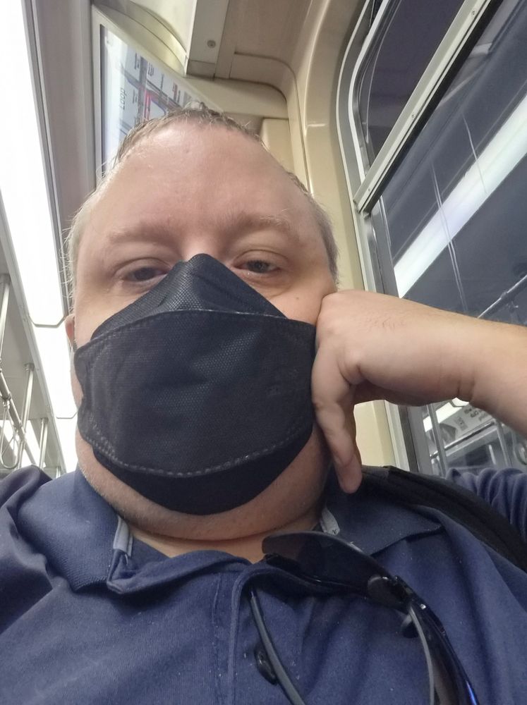 Me, sitting on a train, leaning on my elbow, and wearing a mask over my nose and mouth.