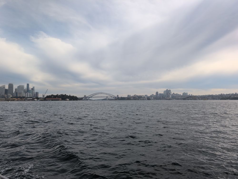 A photo from on Sydney harbour showing the iconic bridge in the distance. The sea is dark and calm and there is a blanket of whispy clouds