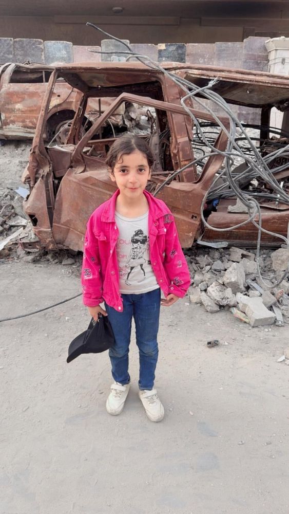 Fidaa’s daughter Layan in front of an exploders car. She wears a pink jacket, white graphic tee and jeans 