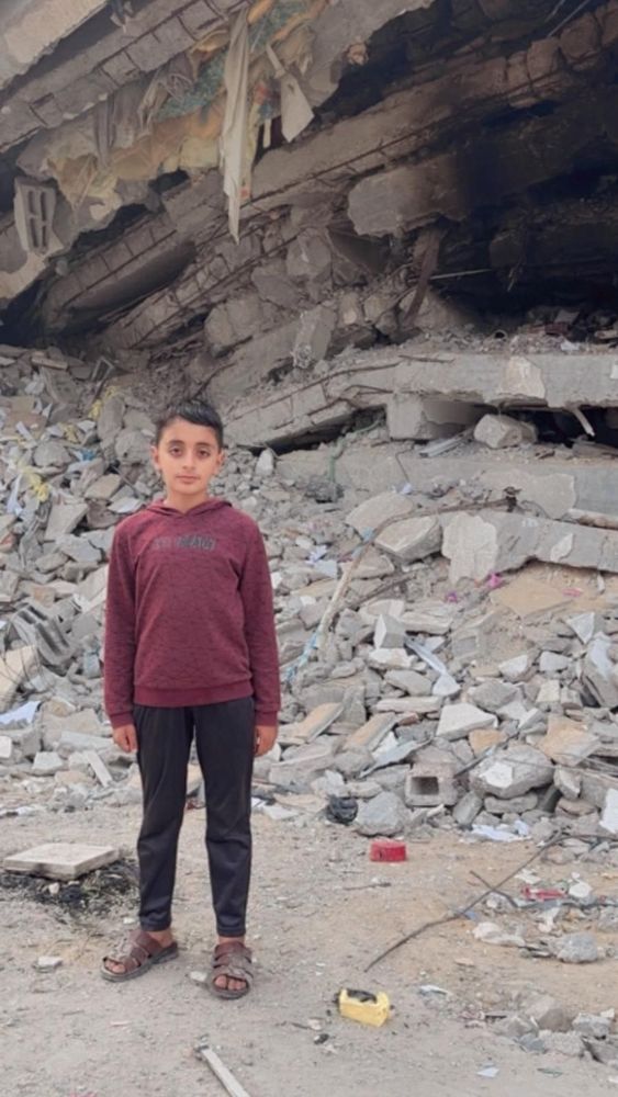 Fidaa’s son Yamen standing in front of rubble. He wears black pants and a brown sweatshirt 