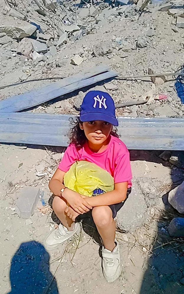 Layan, wearing a blue ball cap, pink shirt, and black shirts, sits on a pile of rubble