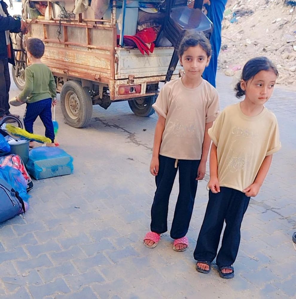 Layan and Mira, wearing t shirts black pants and sandals, stand behind a three-wheel truck with lots of unidentifiable stuff in it 