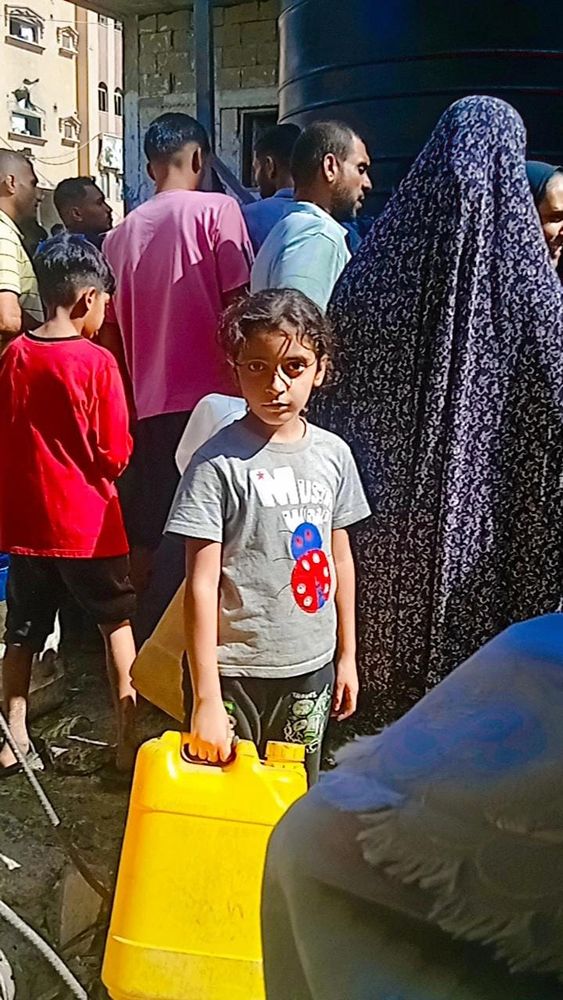 Layan looks at the camera while carrying a large yellow water jug. There are people behind her. She is waiting for water 