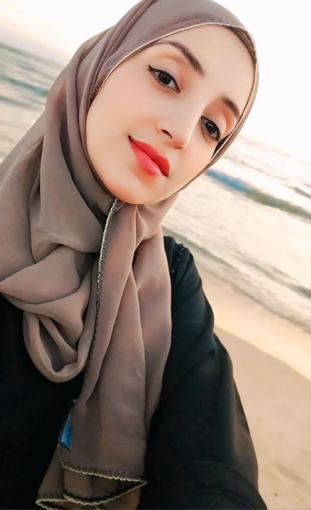 Fidaa takes a selfie on the beach with the waves lapping behind her. She wears a beige hijab and coral lipstick 