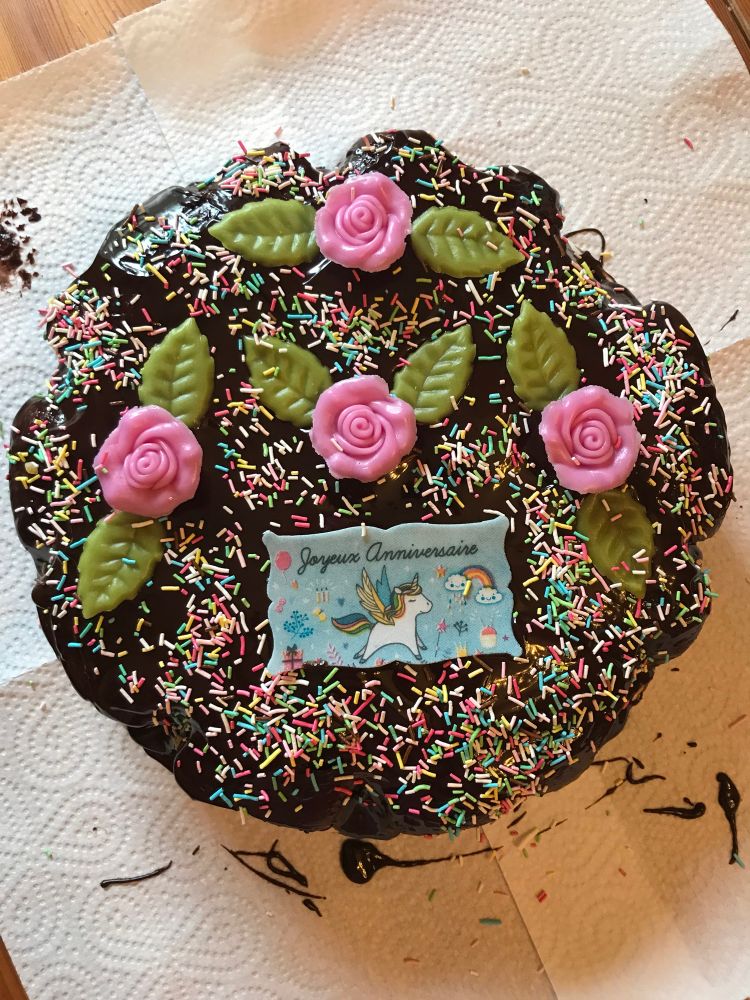 Gâteau au chocolat décoré avec une banderole bleu clair ornée d’une licorne et d’arcs-en-ciel marquée « joyeux anniversaire » , 5 grosses roses rose en sucre et leurs feuilles verte et une multitude de paillettes multicolores 