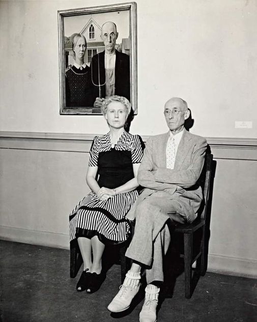 B&w photo of two people sitting in front of Grant Wood’s painting “American Gothic”