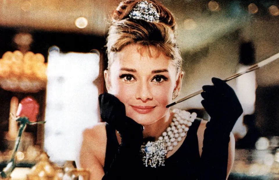 Audrey Hepburn is bejewelled and gloved, elegantly balancing a long cigarette holder, in a still from the film “Breakfast at Tiffany’s.”