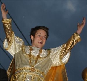 photo of elijah wood in a golden princely costume enthusiastically raising his arms in celebration. it is a cheerful, joyful, and slightly menacing image 