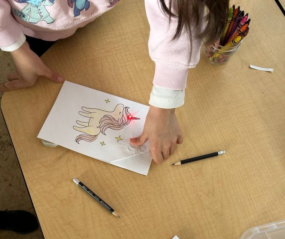 A young student wearing a pink sweater looks down at her light up LED card. There is a yellow unicorn on the front, with a small, red LED in the horn. The student is pressing down on a battery, making the LED light up.