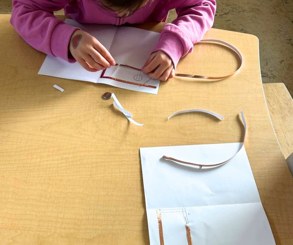 A student wearing a dark pink sweater works on her light up LED card. She is creating a circuit using copper tape.