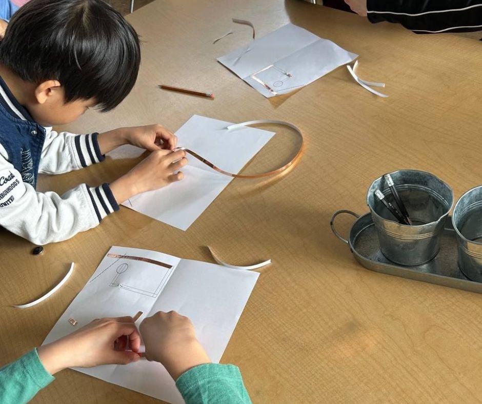 Young students work on creating a light up LED card using paper and copper tape at a wooden table. 
