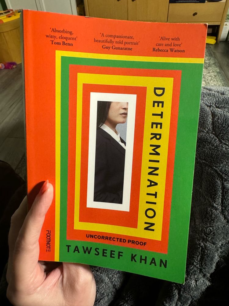 Hand holding a copy of Determination by Tawseef Khan, a striking cover of layered borders in green, orange-red, yellow and white, the half hidden profile of a woman in business attire is at the centre. 