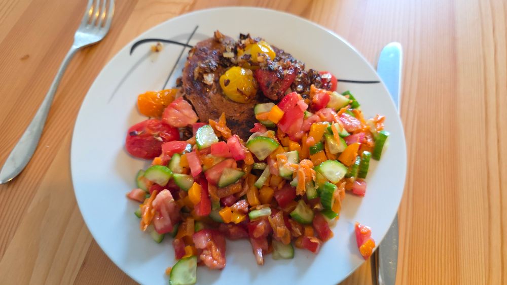 Ein großer weißer Teller steht auf einem Holztisch. Auf dem Teller befindet sich ein vrganes Steak mit angebratenen, fein gehackten, Zwiebeln und Cherry Tomaten.
Daneben ein gemischter Salat aus Tomaten, Gurke, Paprika und Karotte. 